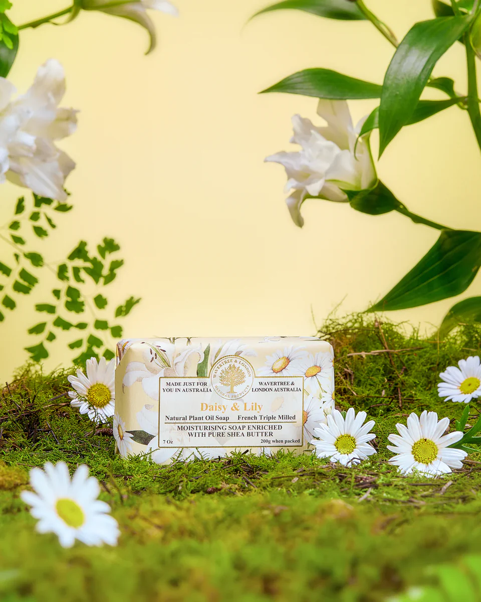 A bar of soap with floral patterns, placed on a bed of moss, surrounded by white daisies and green leaves against a yellow background.
